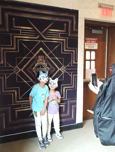 Two children standing in front of a decorative art deco curtain while an adult takes their photo.